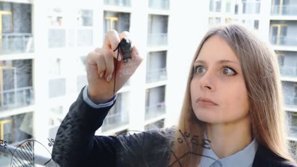 Lifestyle: Schöne junge Frau zeichnet Mathe-Business-Graphen auf das Glas mit Wolkenkratzer im Hintergrund. Nahaufnahme, Handheld, Zeitlupe 60 fps. — Stockvideo