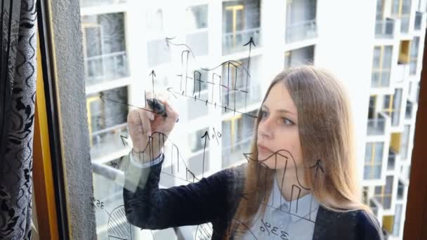 Stile di vita: bella giovane donna che disegna grafici aziendali matematici sul vetro con grattacielo sullo sfondo. Colpo medio, palmare, rallentatore 60 fps . — Video Stock