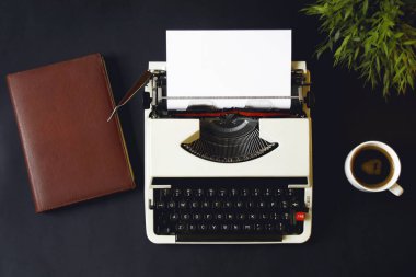 Eski daktilo makinesinin düz yatağı, kırmızı defter, kahve fincanı ve yeşil bitki siyah masada, fotokopi alanı olan kağıt.