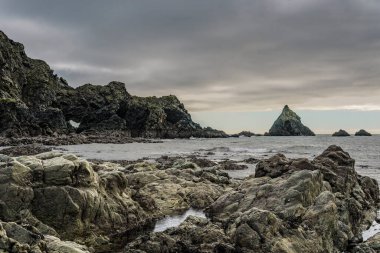 Wild rocky shoreline with sea arch, sea stack, and rocky shore under heavy overcast sky.