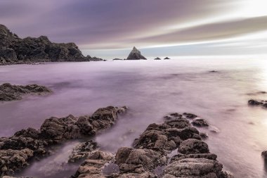Tranquil long-exposure view of Irish coast: silky purple water, textured rocks, and dramatic sea stack.