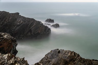 İrlanda kıyılarının uzun süreli deniz manzarası, yumuşak bulutlu ışık altında dalgalı kayaların etrafında dönen ipeksi sisli su..