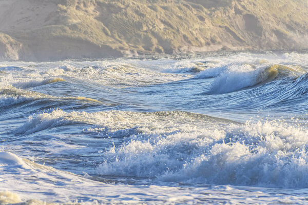 Волны Ирландского моря разбиваются около песчаных дюн, золотой свет подчеркивает пенные гребни и бурную прибрежную воду.