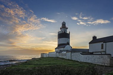 Gün batımında kanca başlı deniz feneri koruyucu evler, engebeli uçurumlar, köpüren Atlantik dalgaları ve parlak kış gökyüzü..