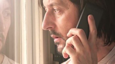 Ciddi adam oturma odası penceresinden dışarı bakıyor ve cep telefonu konuşuyor, yüz görüntüleri yakın.