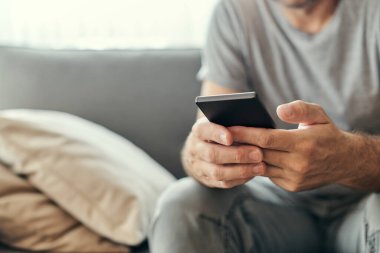 Evde cep telefonu kullanan bir adam, yetişkin beyaz bir erkek oturma odasındaki kanepede sabah rutininin tadını çıkarıyor.