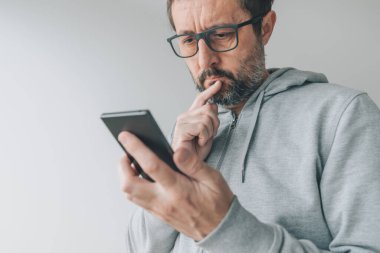 Serious thoughtful mid-adult male looking at mobile smart phone screen with finger on lips and reading text message, close up with selective focus clipart