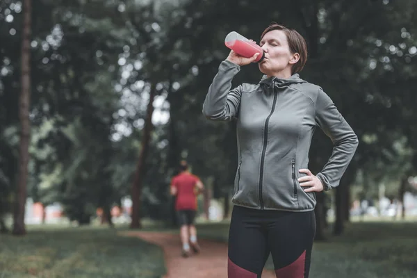 40代の女性ジョガー飲料公園でのさわやかなイチゴジュース 健康的なライフスタイルとレクリエーション活動 — ストック写真