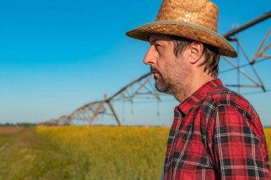 Ciddi bir çiftçinin portresi kolza tohumu tarlasında işlevsiz merkez pivot sulama ekipmanının önünde duruyor, seçici odaklanma