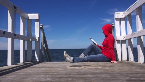 Одинокая женщина в красной рубашке читает на пирсе — стоковое видео