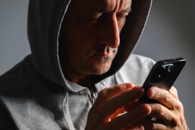 Mature man wearing hoodie using smartphone in low key light, concept of privacy, anonymity, or online communication. Selective focus.