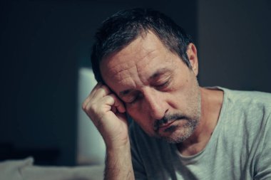 Sleepy tired man resting head on hand with eyes closed in dark low key living room interior at night. Selective focus.