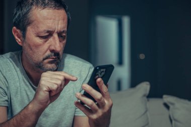Tired mature man typing text message on smartphone in dark living room interior, using mobile phone with index finger, low key portrait in casual home setting. Selective focus.
