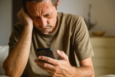 Stressed man holding his head while looking at smartphone, frustrated and worried expression indoors. Selective focus.