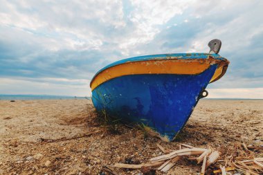 Old fishing boat resting on a sandy beach under a dramatic cloudy sky. The weathered paint and rustic appearance evoke themes of tradition, maritime life, and coastal environments. Selective focus.