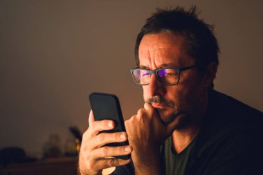 Loş ışıklı odada akıllı telefon kullanan orta yaşlı bir adam gözlüklü mavi ışığın yansımasıyla ekrana odaklanmış. Seçici odak.