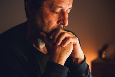 Karanlık odada yalnız başına oturan endişeli bir adam. Sıcak, sade bir ışık, ciddi bir ifade ve duygusal gerilim. Seçici odak.