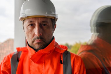 Güvenlik teçhizatındaki düşünceli inşaat mühendisi cama yansıyarak sorumluluğu, planlamayı ve inşaat endüstrisinin insan tarafını sembolize ediyor. Seçici odak.