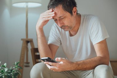 Olgun bir adam akıllı telefonuyla sosyal medyada oyalanıyor ve geziniyor. Yorgun ve sıkılmış bir şekilde oturma odasındaki rahat koltukta, lambanın yanında yatıyor. Seçici odak.