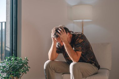 Depresyonda olan bir adam ellerini başının üstüne koyup öne eğiyor. Parlak ve sessiz bir oturma odasında endişe ve kötü haberlerle boğuşuyor. Seçici odak.