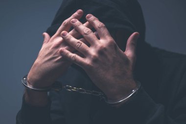 Kapüşonlu bir suçlunun göze batmayan portresi. Elleri yüzü kapalı, elleri kelepçeli. Görünür ve dramatik gölgeler gerginliği ve suçluluğu vurguluyor. Seçici odak.