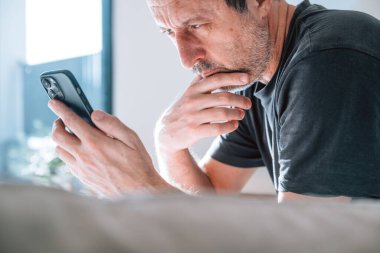 Otantik orta yaşlı beyaz bir adam, eli çenesinde, akıllı telefona bakarken son derece endişeli görünüyor. Pencereden gelen yumuşak doğal gün ışığı. Kötü haber, borç, stres, endişe ve kriz için güçlü bir kavram