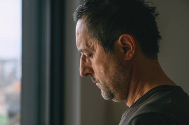 Pencere kenarında duran olgun bir adamın portresi, yıpranmış, yorgun bir ifadeyle, dağınık saçlı ve sakallı, yorgunluk ve duygusal ağırlığı gösteren bir portre. Seçici odak.