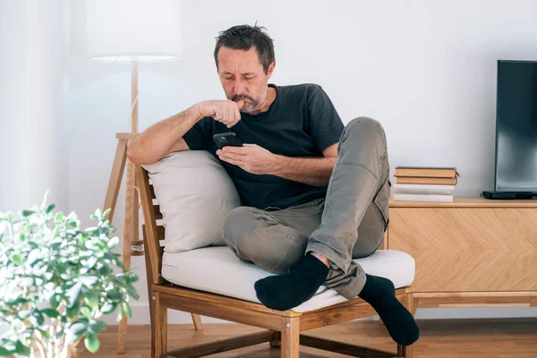 Yorgun, orta yaşlı bir adam güneş ışığıyla aydınlanmış bir koltuğun derinliklerine iniyor, siyah çoraplarla bacak bacak bacak bacak üstüne atıyor, telefonunu kapatırken gözleri sessiz bir alarmda daralıyor. Parlak, minimalist bir oturma odasında saf, samimi bir modern endişe anı..