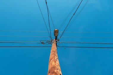 Yıpranmış ahşap elektrik direği, karışık elektrik kabloları seramik yalıtıcılar ve parlak mavi gökyüzüne karşı eski bir sokak lambası. Seçici odak.
