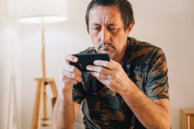 Odaklanmış orta yaşlı bir adam, akıllı telefonu üzerinde çalışırken öne doğru eğilir, yüzü güneş ışığıyla gerilir ve odayı aydınlatır. Seçici odak.