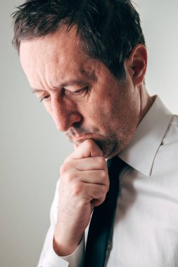 Beyaz gömlekli ve koyu renk kravatlı bir iş adamının portresi, elleri çene üzerinde düşünceli bir şekilde dinlenirken aşağıya, seçici bir şekilde odaklanıyor.