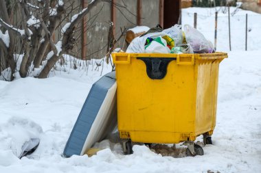 Karlı kış bahçesinde çöp torbalarıyla dolup taşan sarı çöp konteynırı, seçici bir odak noktası