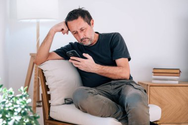 Hakiki orta yaşlı beyaz adam, akıllı telefondan kayarken yorgun ve bitkin görünüyor. Zihinsel yorgunluk, yorgunluk, sosyal medya bağımlılığı, ruh sağlığı teması. Seçici odak.