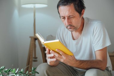 Sıradan, dağınık saçlı, rahat bir oturma odasında sarı kaplı bir kitap okuyan, sabah ışığında rahat bir ev atmosferi yaratan olgun bir adam. Seçici odak.