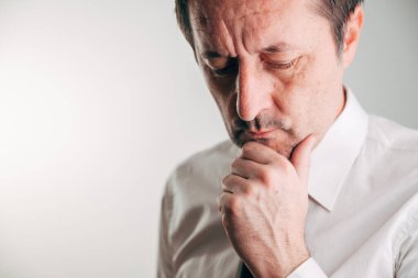 Beyaz gömlekli ve kravatlı orta yaşlı bir işadamı, elleri çenesinde aşağıya bakıyor, stres, endişe ve derin düşüncelerini tarafsız bir arkaplanda ifade ediyor. Seçici odak.