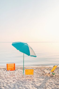 Renkli şezlonglar ve yumuşak kumda turkuaz şemsiyeli, pastel bir gündoğumu gökyüzünün altında sakin deniz sularına bakan minimalist yaz plajı sahnesi. Huzurlu bir tatil ortamı. Seçici odak.