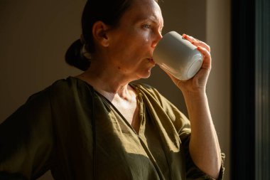 Orta yaşlı bir kadının kahve ya da çay içtiği, altın güneş ışığının yumuşak jant ışığı yarattığı ve sakin yansıtıcı ruh hali. Seçici odak.