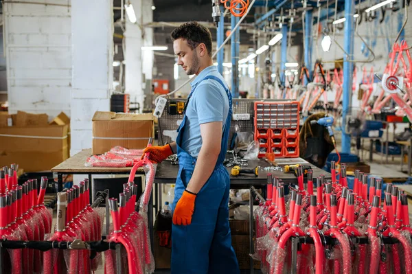 Bicycle factory, worker holds pink kids bike fork. Male mechanic in uniform installs cycle parts, assembly line in workshop, industrial manufacturing