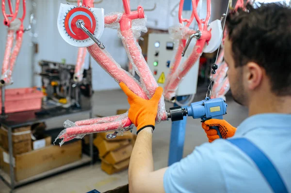 Bicycle factory, worker holds pink kids bike. Male mechanic in uniform installs cycle parts, assembly line in workshop, industrial manufacturing