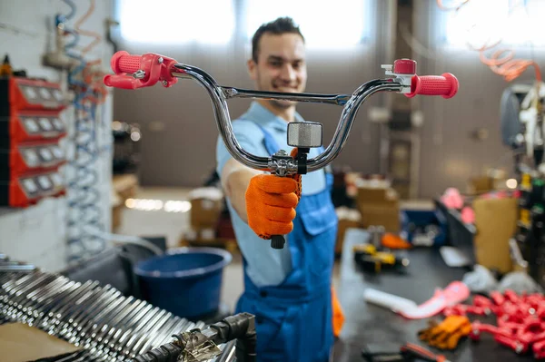 Bicycle factory, worker shows girls bike handlebar. Male mechanic in uniform installs cycle parts, assembly line in workshop, industrial manufacturing