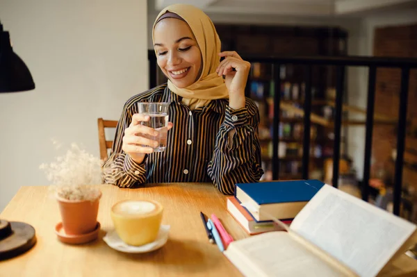 Arab girl in hijab holds glass of water, university cafe interior on background. Muslim woman with books sitting in library. Religion and education