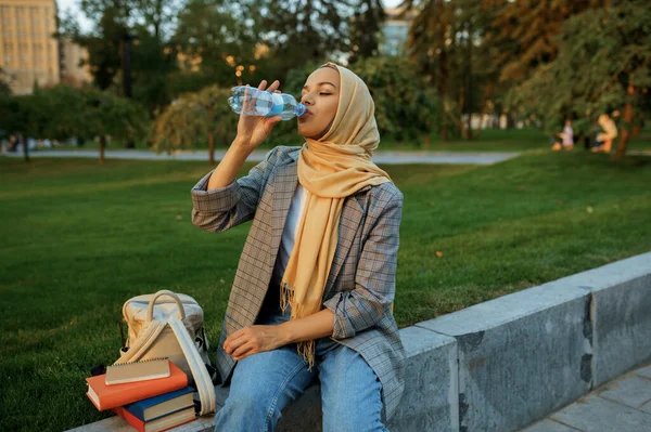 夏の公園でヒジャーブの女性学生は水を飲む 本が芝生の上で休んでいるイスラム教徒の女性 宗教と教育 — ストック写真