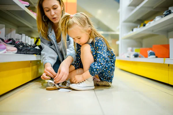 Mother and little baby trying on shoes in kids store. Mom and adorable girl near the showcase in childrens shop, happy childhood, family makes a purchase in market