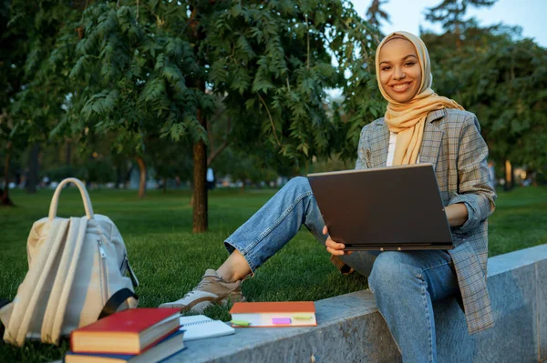 Arab female student in hijab using laptop in summer park. Muslim woman with books resting on the lawn. Religion and education