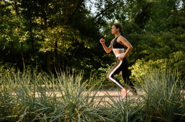 Sabah koşusu, yaz parkında eğitim gören bir kadın. Kadın koşucu güneşli bir günde spor için koşar, sağlıklı bir yaşam tarzı, açık havada koşar.