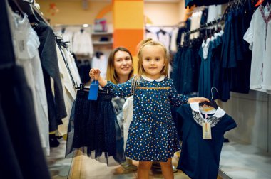 Anne ve küçük bebek çocuk mağazasında etek seçiyorlar. Anne ve sevimli kız çocuk dükkanında vitrinin yanında, mutlu çocukluk, aile çocuk pazarından alışveriş yapıyor.