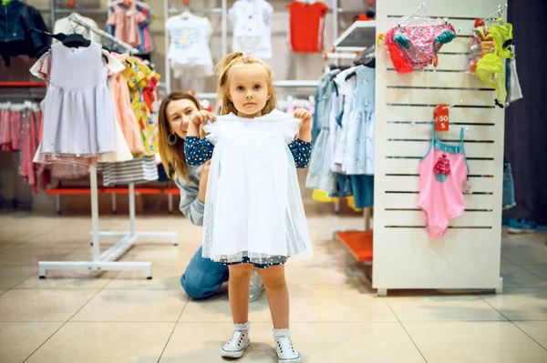 母親と小さな赤ちゃんが子供の店でドレスを試着しています 子供の店でショーケースの近くのお母さんと愛らしい女の子 幸せな子供時代 家族は市場で購入します — ストック写真