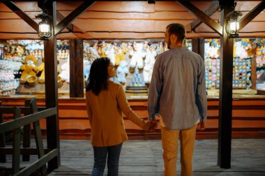 Lunaparktaki hediyelik eşya dükkanında aşk çifti. Erkek ve kadın dışarıda, panayır arka planda. Atlıkarıncalarda aile eğlenceleri, eğlence teması