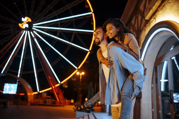 Love couple walking in night amusement park, piggyback ride. Man and woman relax outdoors, ferris wheel with lights on background. Family leisures on carousels, entertainment theme