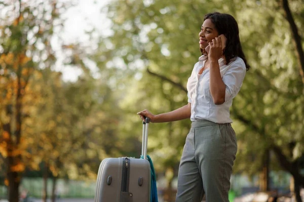 带着手提箱的年轻女人在公园里通过电话交谈 携带行李的女旅客在户外休息 携带行李的旅客在大自然中休息 带着行李的女孩在市区小巷里休息 — 图库照片